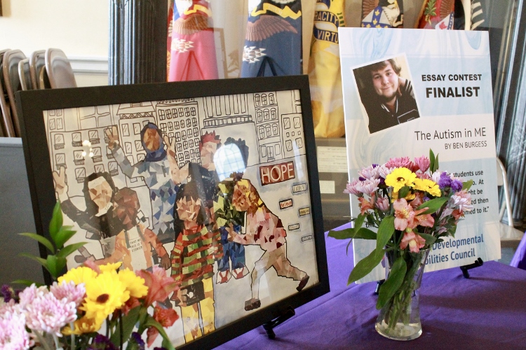 Display of artwork, a poster highlighting one of the essay winners, and vases of flowers at the Inclusion Ceremony